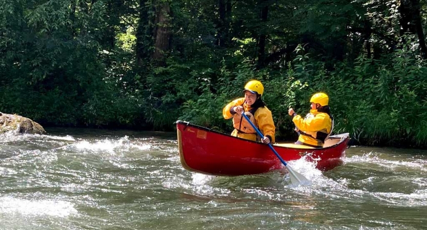 Two people wearing safety gear paddle a canoe beside a shore with thick greenery 